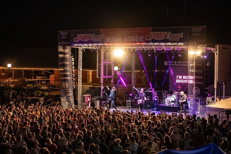 A band performs on a brightly lit outdoor stage at night before a large, cheering crowd at a music festival.
