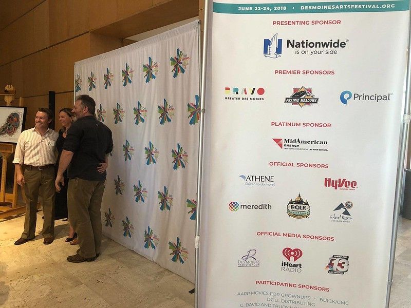 A vertical banner listing event sponsors next to two people standing in front of a patterned backdrop in an indoor space.