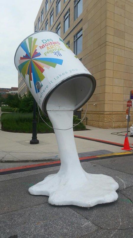 A large, sculptural paint bucket tilted on its side with white paint spilling out onto the sidewalk, promoting an art fest.