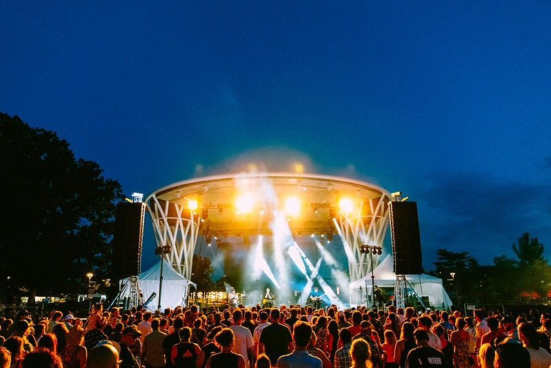 A large crowd faces a brightly lit outdoor stage under a twilight blue sky, with beams of light shining into the night.