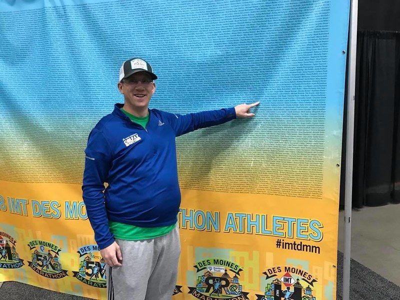 A person in a blue zip-up and baseball cap smiles while pointing to their name on a list of marathon athletes.