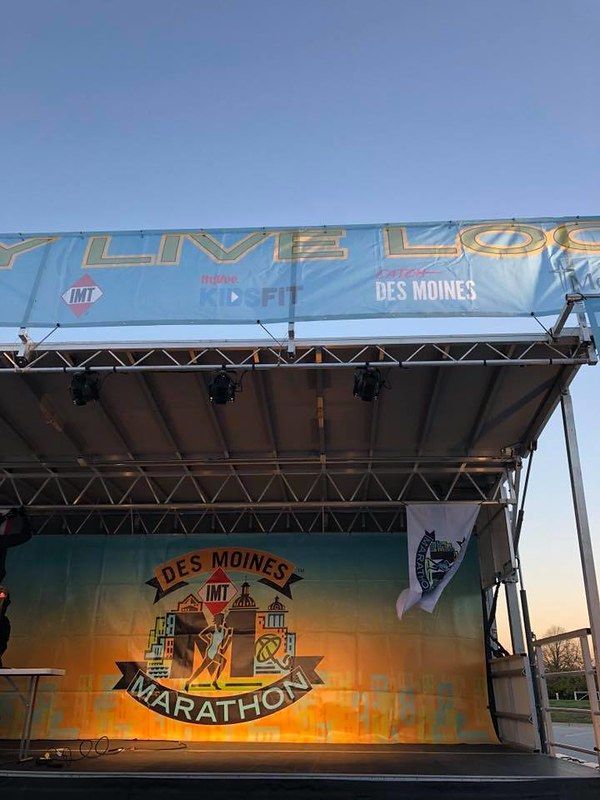 An outdoor stage for the Des Moines Marathon, featuring event signage and branding against a sunset-colored backdrop.