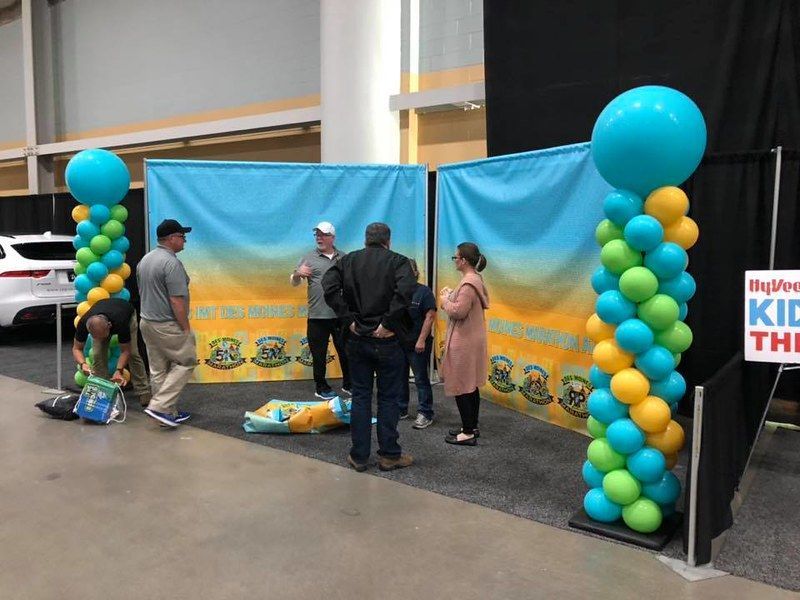 Four people stand in front of two light blue and yellow display banners, framed by two vertical blue and green balloon towers.