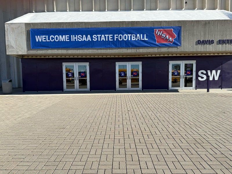 A banner welcoming guests to the IHSAA State Football championships hangs above three glass entrance doors on a building.