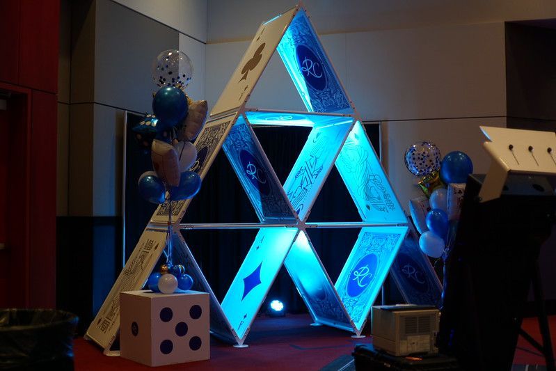 A large, light-up sculpture of a house of cards with blue glowing panels, accented by blue and silver balloons and a die.