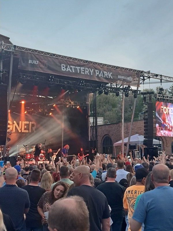 A live rock concert at Battery Park with a band performing on stage before a large, cheering crowd.