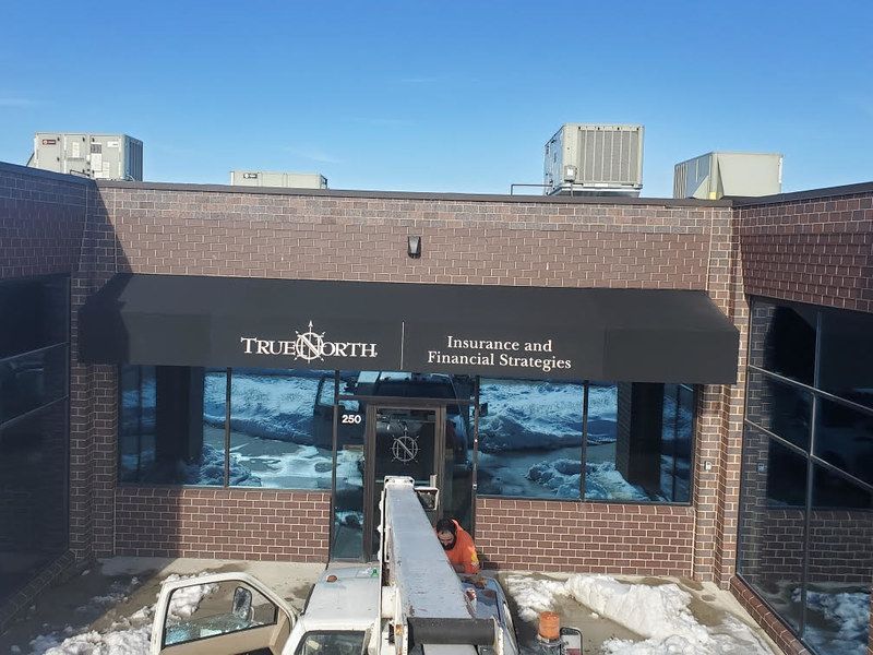 A worker in an orange vest uses a lift to install a black awning on a brick building for True North Financial Strategies.
