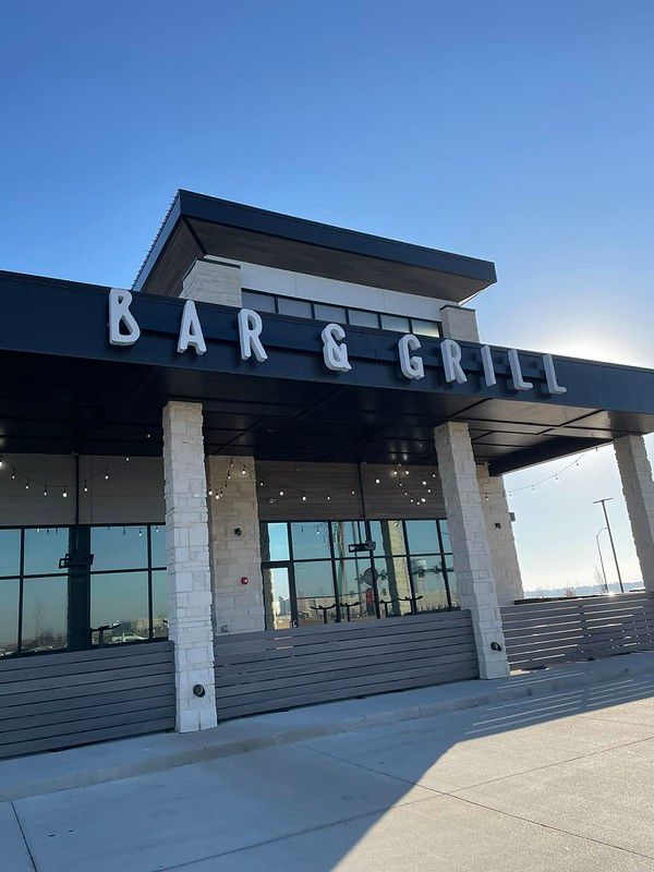 Modern building exterior with large glass windows and stone pillars, featuring a prominent “BAR & GRILL” sign above.