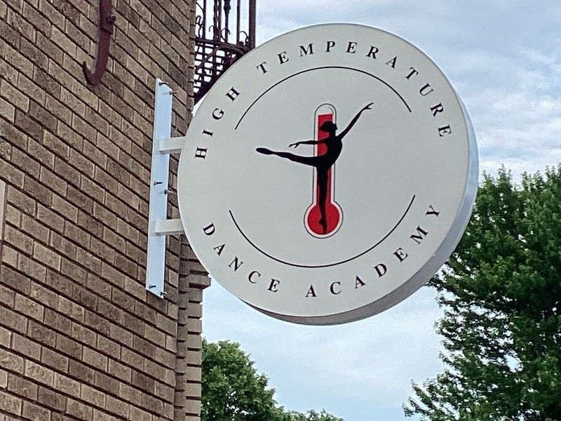 A circular, white sign for High Temperature Dance Academy featuring a thermometer with a dancer silhouette, on a brick wall.
