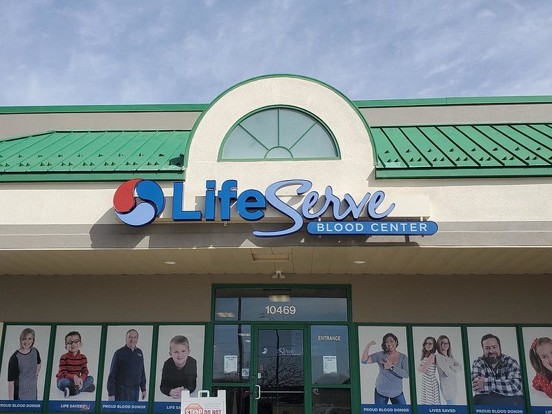 The exterior of a LifeServe Blood Center with a green roof, blue signage, and photos of people displayed on the windows.