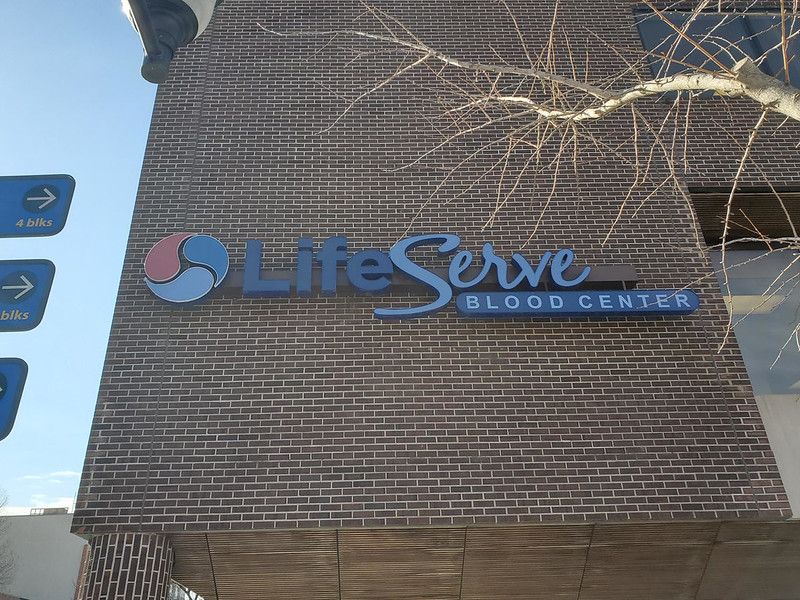 A LifeServe Blood Center sign mounted on a dark brick building exterior under a bright sky.