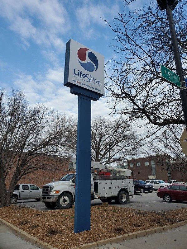 A LifeSprout sign stands on a blue pole in a parking lot with trees, a utility truck, and other cars in the background.