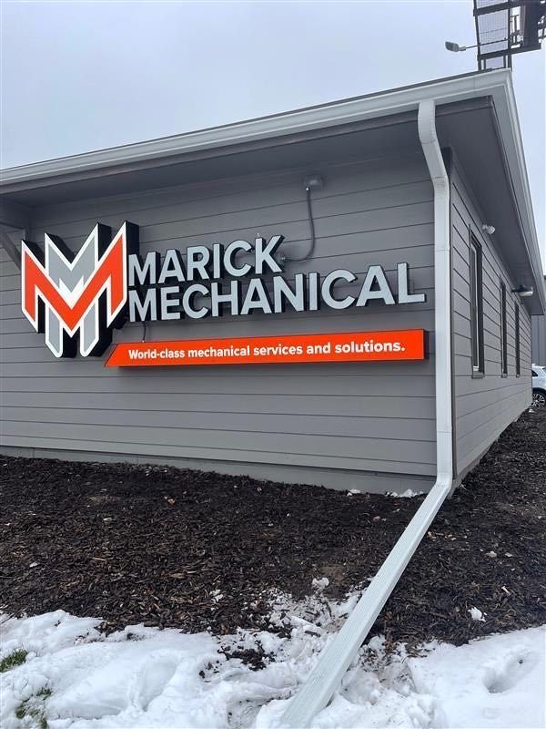 Sign for Marick Mechanical on the exterior of a gray building, featuring an orange and gray logo on a bed of dark mulch.