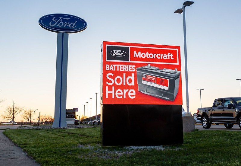 A Ford dealership sign in a grassy lot advertises Motorcraft batteries for sale, with a tall Ford logo pole in the back.