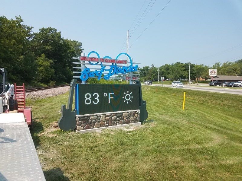 A welcome sign for Clear Lake, Iowa, shaped like a guitar, displaying an 83°F temperature on a sunny day.