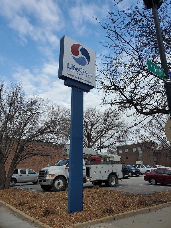 A LifeSpire sign stands on a blue pole in a parking lot with trees and vehicles under a partly cloudy sky.