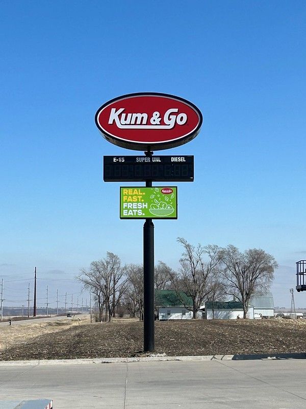 A roadside Kum & Go gas station sign against a clear blue sky, featuring a red oval logo and a green digital display.