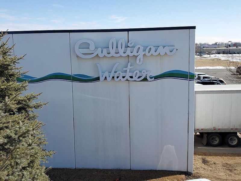 A Culligan Water sign mounted on a white wall outdoors next to a evergreen tree and a parked semi-trailer.