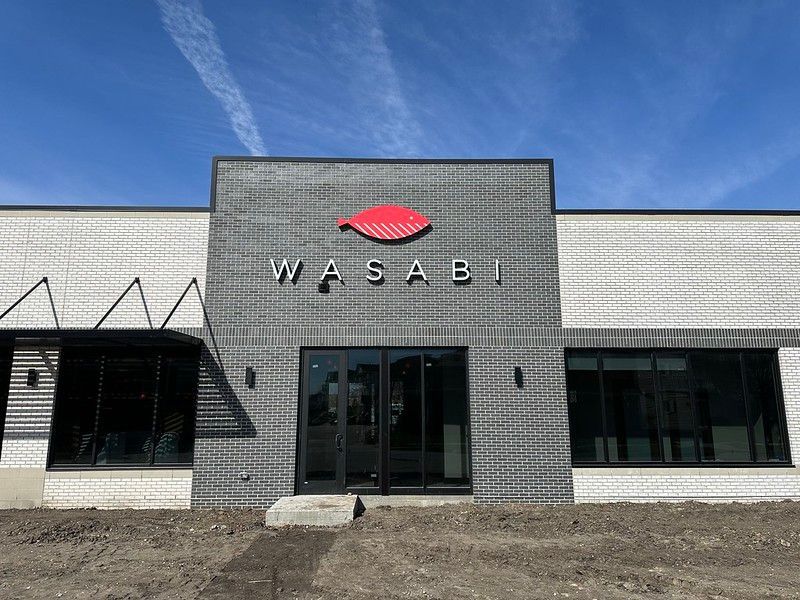Exterior of the Wasabi restaurant featuring a grey brick facade, a red leaf logo, and large black-framed windows.