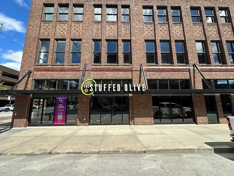 The Stuffed Olive restaurant storefront in a multi-story brick building under a clear blue sky.