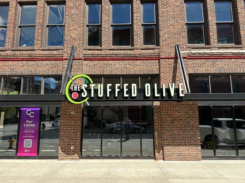 Storefront of The Stuffed Olive with a brick facade, floor-to-ceiling windows, and a For Lease sign on the left.