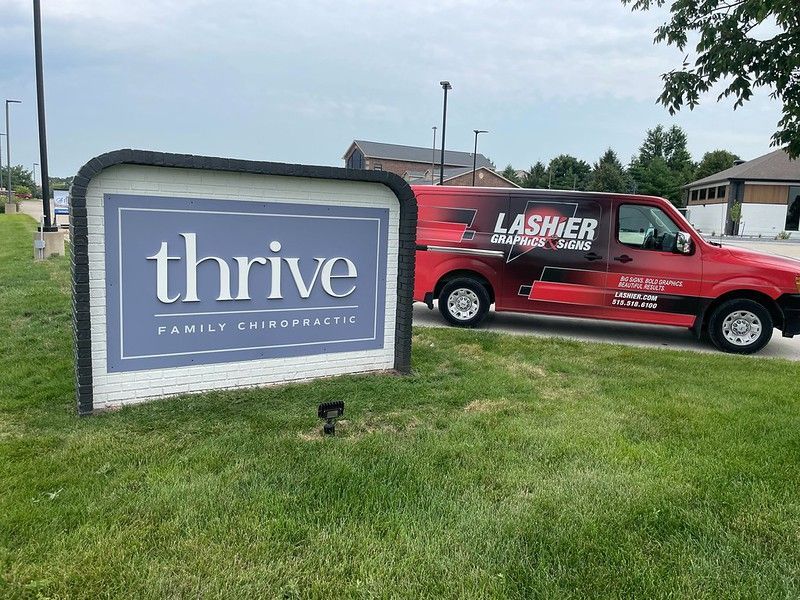 A sign for Thrive Family Chiropractic stands in a grassy lawn next to a red Lashier Landscaping van.