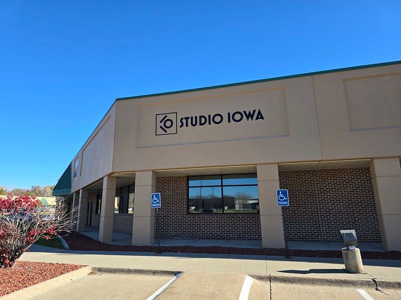 A tan building with the Studio Iowa logo on the exterior, featuring a sidewalk and two handicap parking signs.