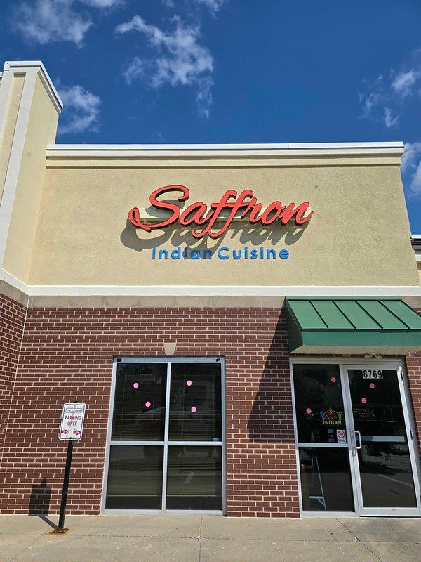 The storefront of Saffron Indian Cuisine, featuring a beige upper wall, red and blue signage, and brick exterior.