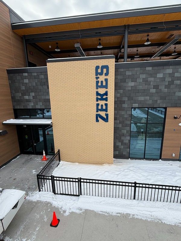 Exterior view of Zeke’s restaurant with blue signage on a tan brick wall, dark stone siding, glass doors, and snowy ground.
