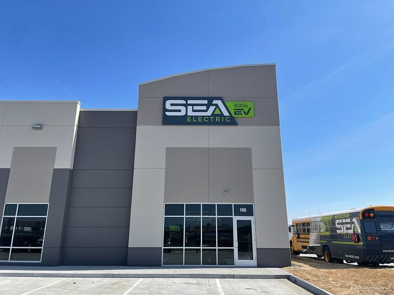 Exterior of a Sea Electric building with a company-branded school bus parked nearby under a clear blue sky.