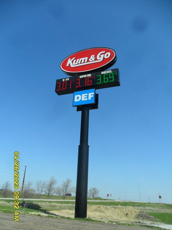 A tall Kum & Go gas station sign against a clear blue sky, displaying prices for various fuels.