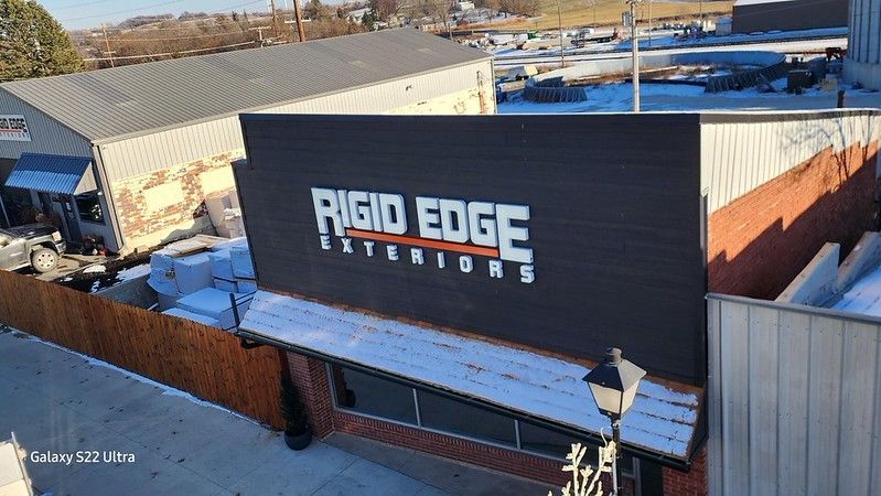 A high-angle view of the black Rigid Edge Exteriors sign mounted on a brick building during a winter day.