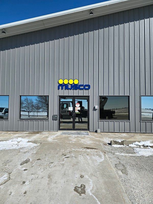A grey metal industrial building with a blue Musco logo and yellow circles above the glass entrance doors.