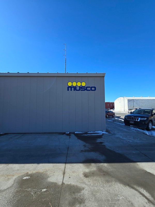 A gray industrial building features a Musco company logo with yellow circles above blue text under a clear blue sky.