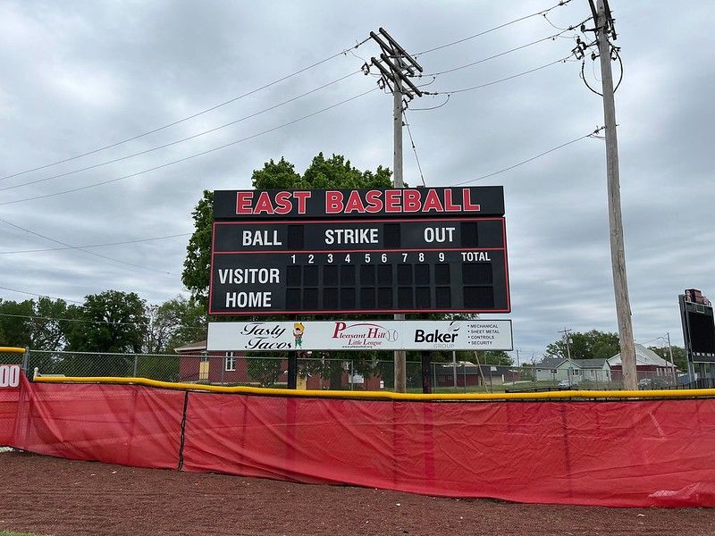 A baseball scoreboard at East Baseball stadium featuring innings and game stats, set behind a red outfield fence.