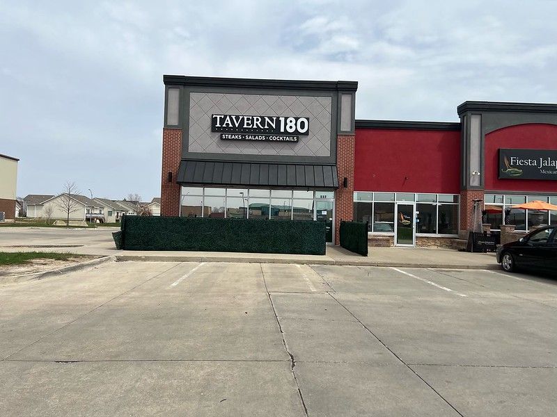 Tavern 180 restaurant exterior with a grey brick facade, dark awning, and a green privacy screen in a parking lot.