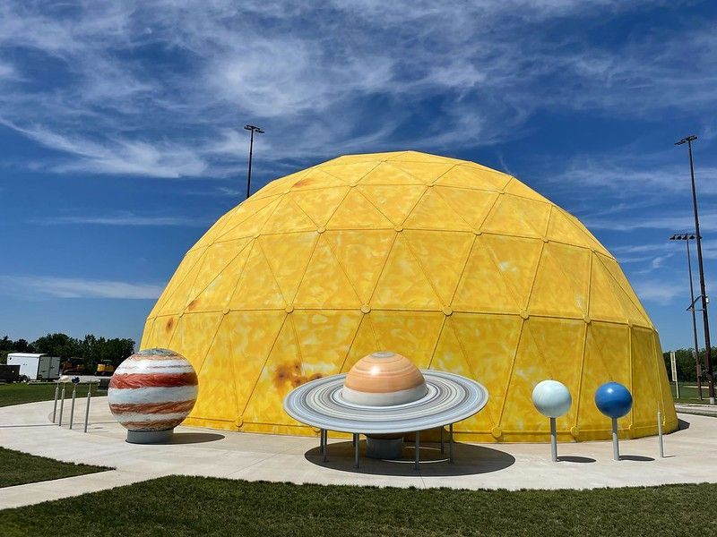A large yellow geodesic dome resembling the sun, with models of planets positioned in front against a blue sky.