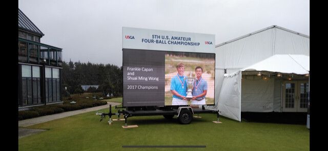 A mobile digital billboard outdoors displays two people holding a trophy, identifying them as 2021 U.S. Four-Ball champions.