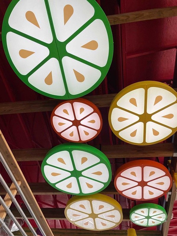 Circular citrus slice-shaped lights in shades of green, orange, and yellow hanging from a ceiling with wooden beams.