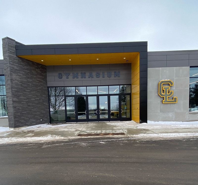 Exterior of a school gymnasium building with grey stone accents, a yellow overhang, and a gold “CL” logo on the wall.