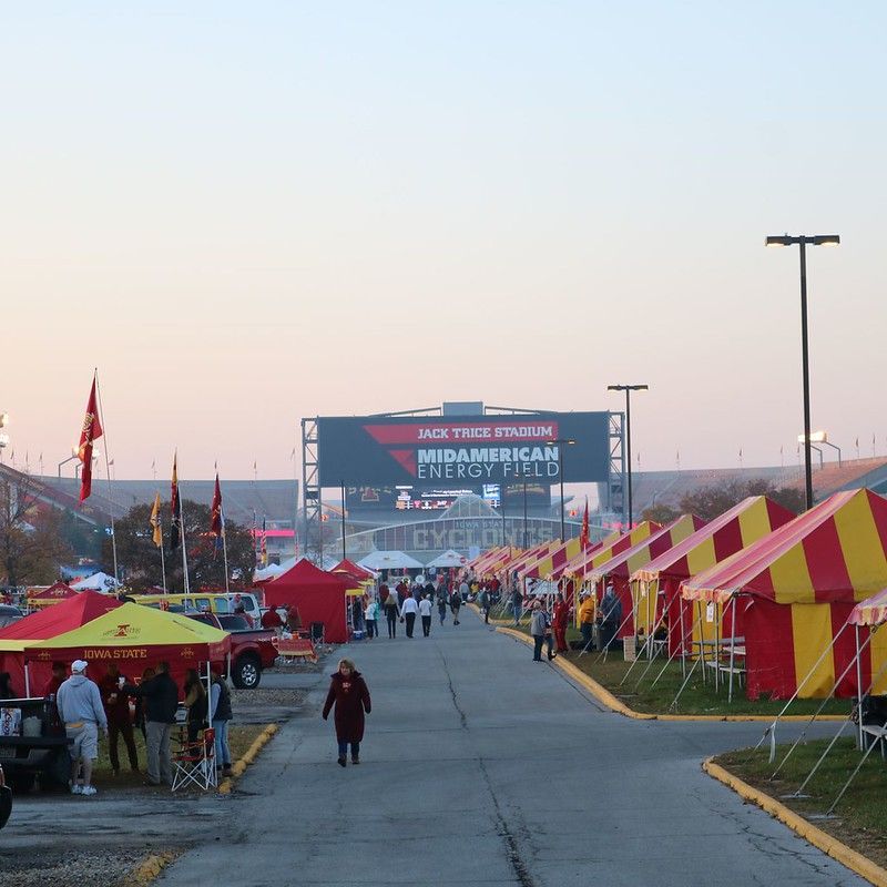 Rows of red and yellow tailgating tents line a paved road leading to a stadium under a pale sky.