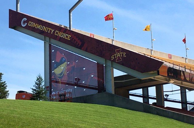 Exterior view of a stadium corner with a cardinal logo, Community Choice branding, and Iowa State flags against blue sky.