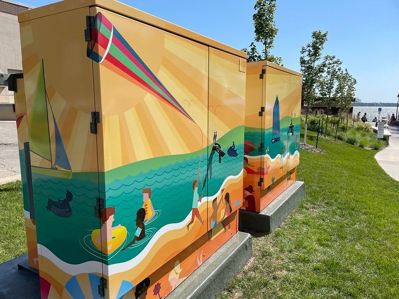 Two utility boxes painted with a colorful beach mural showing people playing in the water and a sailboat under a kite.