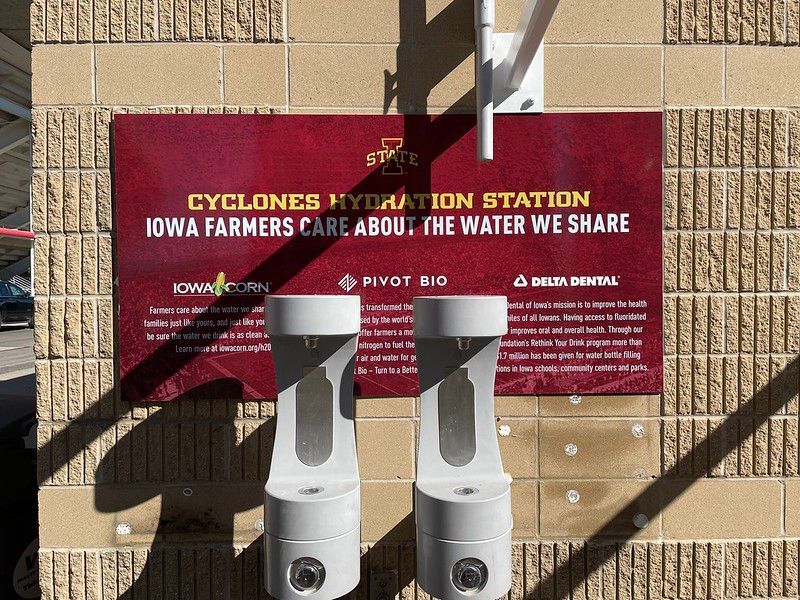 Two water bottle filling stations mounted on a brick wall beneath a maroon sign promoting Iowa farmers and water quality.