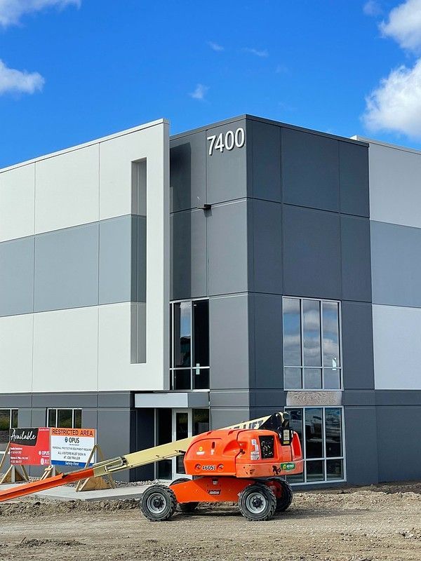 A modern commercial building with gray and white panels, numbered 7400, features an orange construction lift in front.