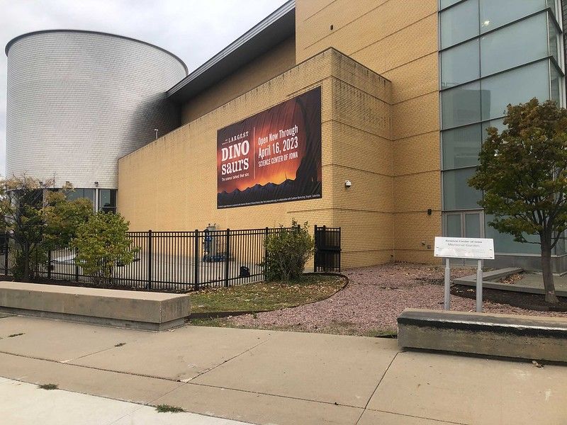 An exterior view of a building with a large DINO SAFARI sign, a cylindrical metal structure, and a sidewalk in front.