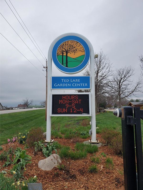 A sign for Ted Lare Garden Center with a circular tree logo and an LED display listing business hours.