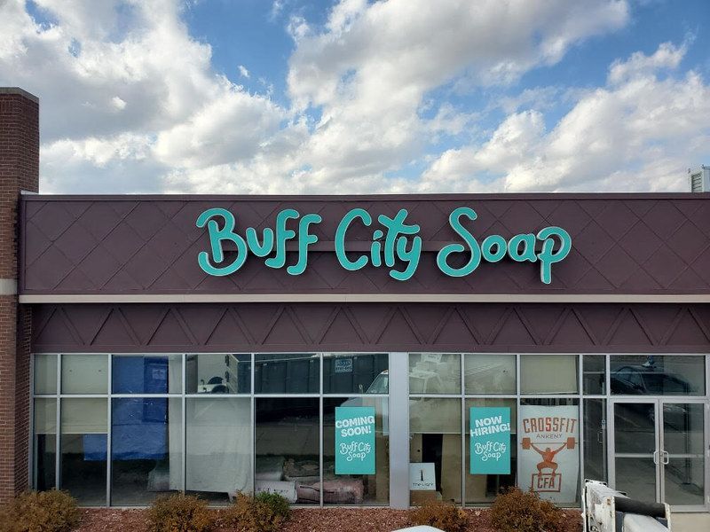 Storefront of a Buff City Soap shop with teal, script-style signage on a dark brown facade against a partly cloudy sky.