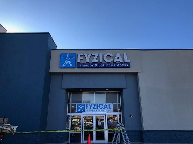 Exterior of a Fyzical Therapy & Balance Centers building under a clear blue sky, with a ladder near the glass entrance.
