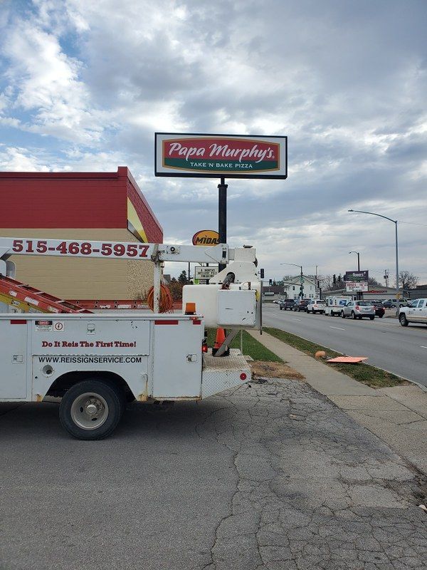 A Papa Murphy's pizza sign stands above a utility truck parked in a lot along a street on a cloudy day.